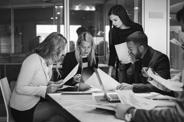 Black and white collage of 5 different photos. Person pointing to someone's laptop, a colleague delivering a presentation to their peers, people raising their hands to ask questions to the speaker, a work place with several people smiling and a focused image of a woman smiling.